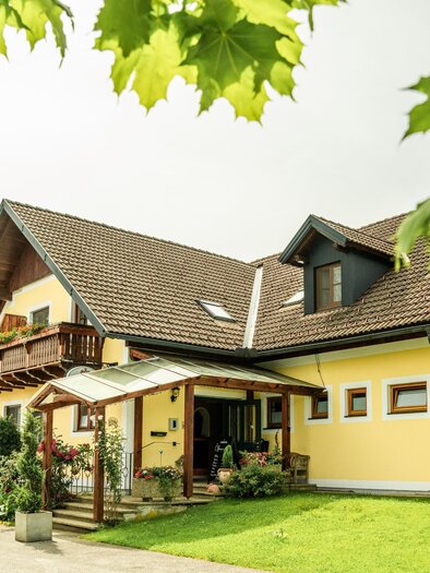 Landhof Lazarus in Mooskirchen mit gelber Fassade, Holzbalkon, überdachtem Eingang und Garten | © Region Graz - Mias Photoart