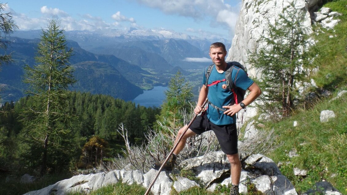 Ein Mann steht auf einem felsigen Weg in den Bergen und hält einen Wanderstock. Im Hintergrund sind grüne Wälder und ein klarer See mit Bergen in der Ferne zu sehen. | © Gerhard Weissenbacher