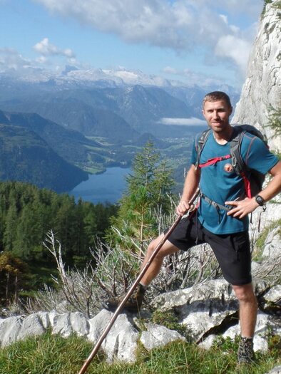 Ein Mann steht auf einem felsigen Weg in den Bergen und hält einen Wanderstock. Im Hintergrund sind grüne Wälder und ein klarer See mit Bergen in der Ferne zu sehen. | © Gerhard Weissenbacher