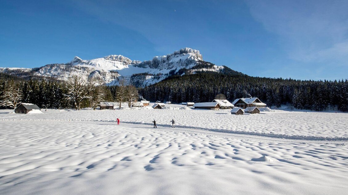 Eine verschneite Landschaft mit mehreren Hütten und Bäumen. Im Hintergrund erhebt sich ein imposantes Gebirge unter einem klaren blauen Himmel. | © TVB Ausseerland - Salzkammergut-Tom Lamm