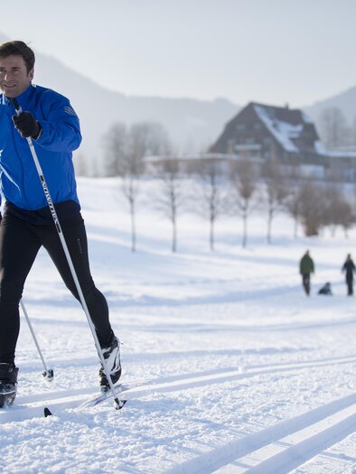 <p>A man in a blue ski jacket is cross-country skiing on the trail in Bad Aussee. In the background, the white winter landscape of Ausseerland with the Hotel Die Wasnerin can be seen in the distance.</p> | © Christian Jungwirth