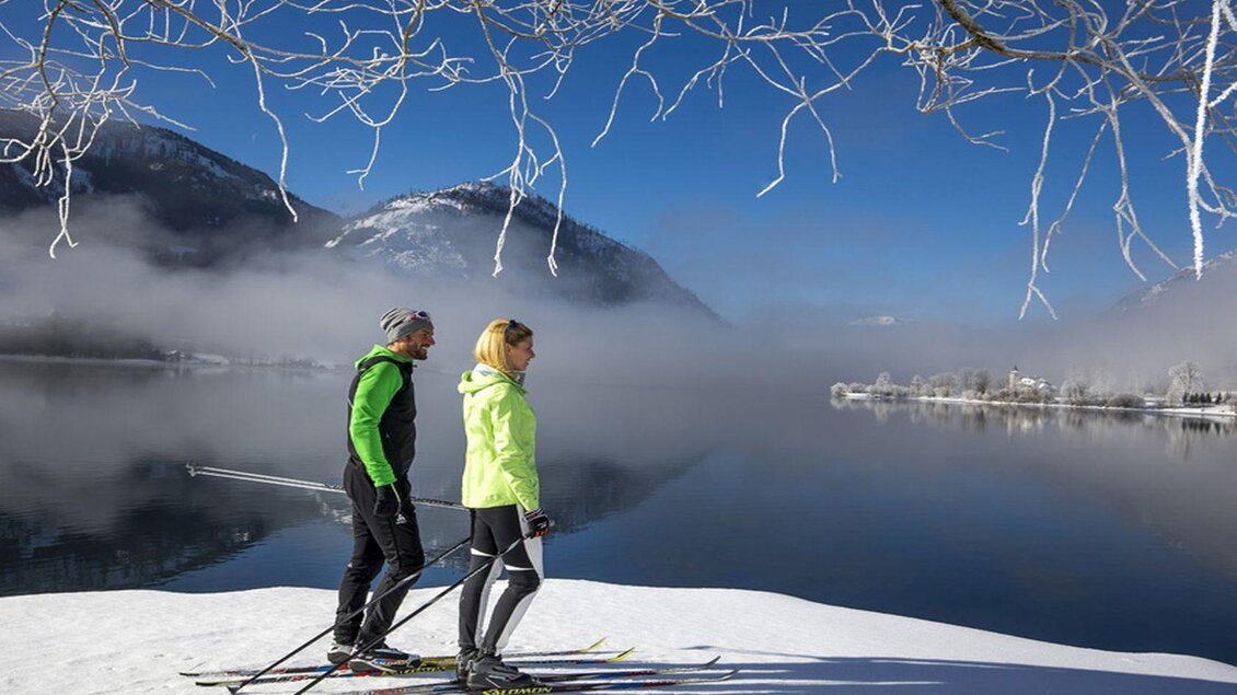 <p>Zwei Personen genießen die Aussicht auf den Grundlsee nach dem Langlaufen in Gößl am schneebedeckten Ufer. Im Hintergrund spiegelt sich der klare Himmel im ruhigen Wasser des Sees.</p> | © TVB Ausseerland - Salzkammergut/Tom Lamm