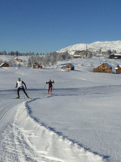 <p><p>Two cross-country skiers are moving through the snowy winter landscape of Tauplitzalm. In the background, some huts and mountains are visible.</p></p> | © TVB Ausseerland Salzkammergut/Fuchs