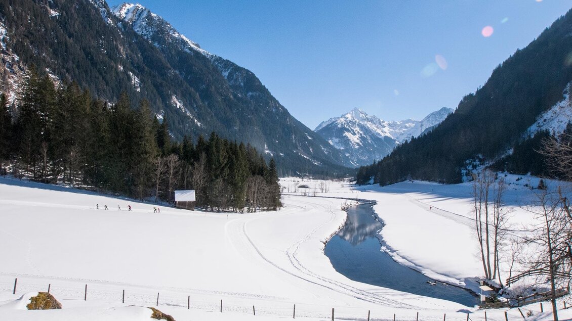 Eine verschneite Landschaft mit hohen Bergen und einem ruhigen Fluss. Im Hintergrund sind Nadelbäume und ein klarer blauer Himmel zu sehen. | © Gerhard Pilz/Gerhard Pilz