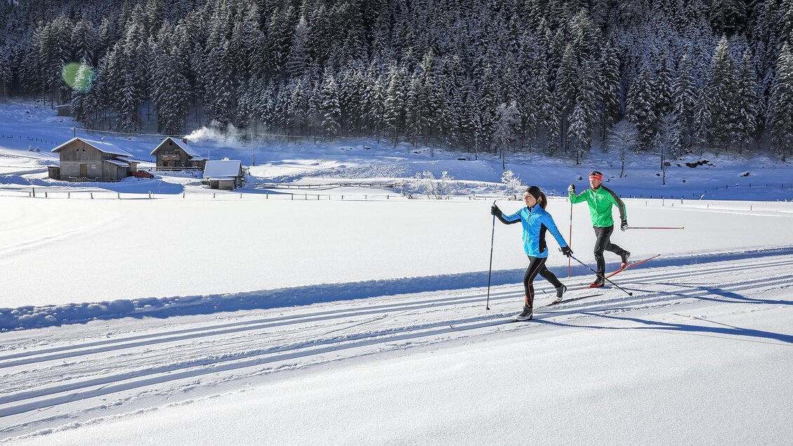 Zwei Personen wandern mit Langlaufskiern durch eine schneebedeckte Landschaft. Im Hintergrund sind verschneite Bäume und einige Hütten zu sehen. | © Gerhard Pilz/Tourismusverband Schladming - Martin Huber