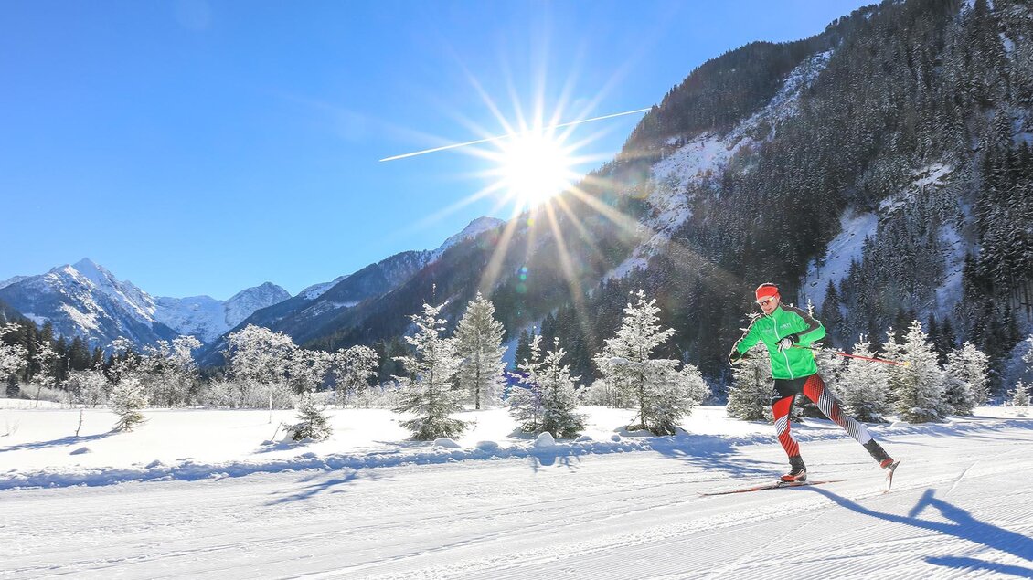 Eine Person fährt Ski in einer verschneiten Landschaft. Im Hintergrund sind Berge und ein strahlender Sonnenschein zu sehen.