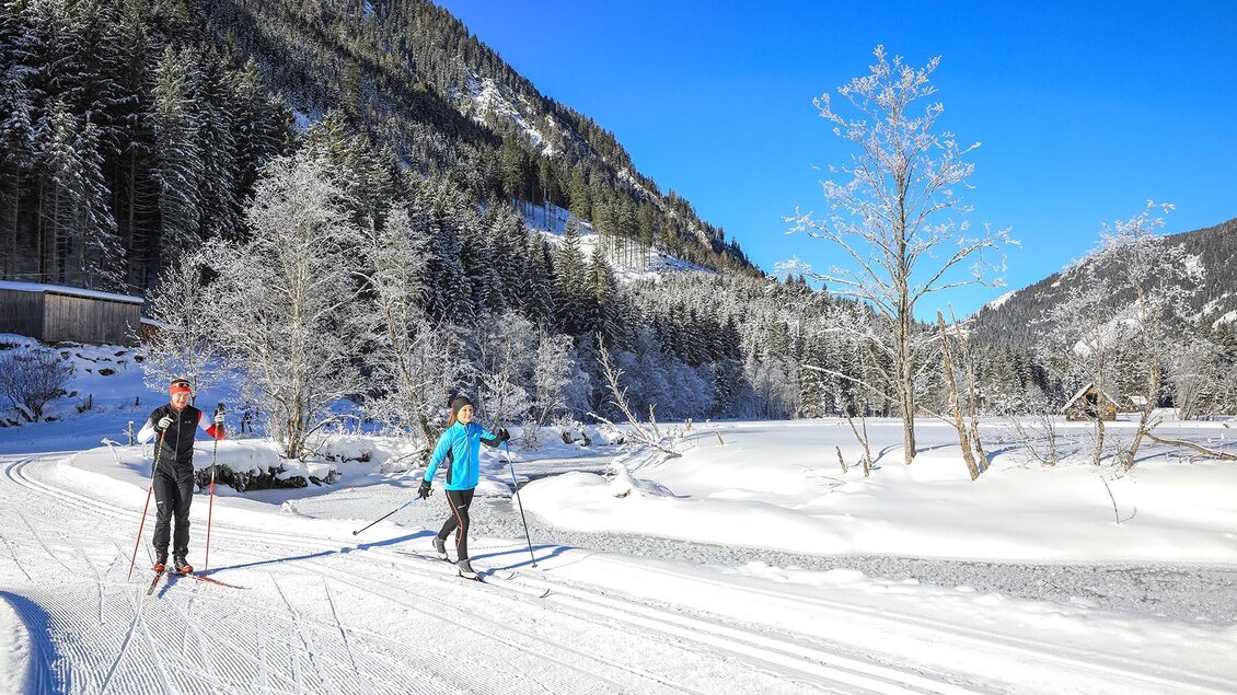 Zwei Menschen beim Langlaufen auf einer schneebedeckten Strecke. Im Hintergrund sind verschneite Bäume und ein blauer Himmel zu sehen. | © Gerhard Pilz/Tourismusverband Schladming - Martin Huber