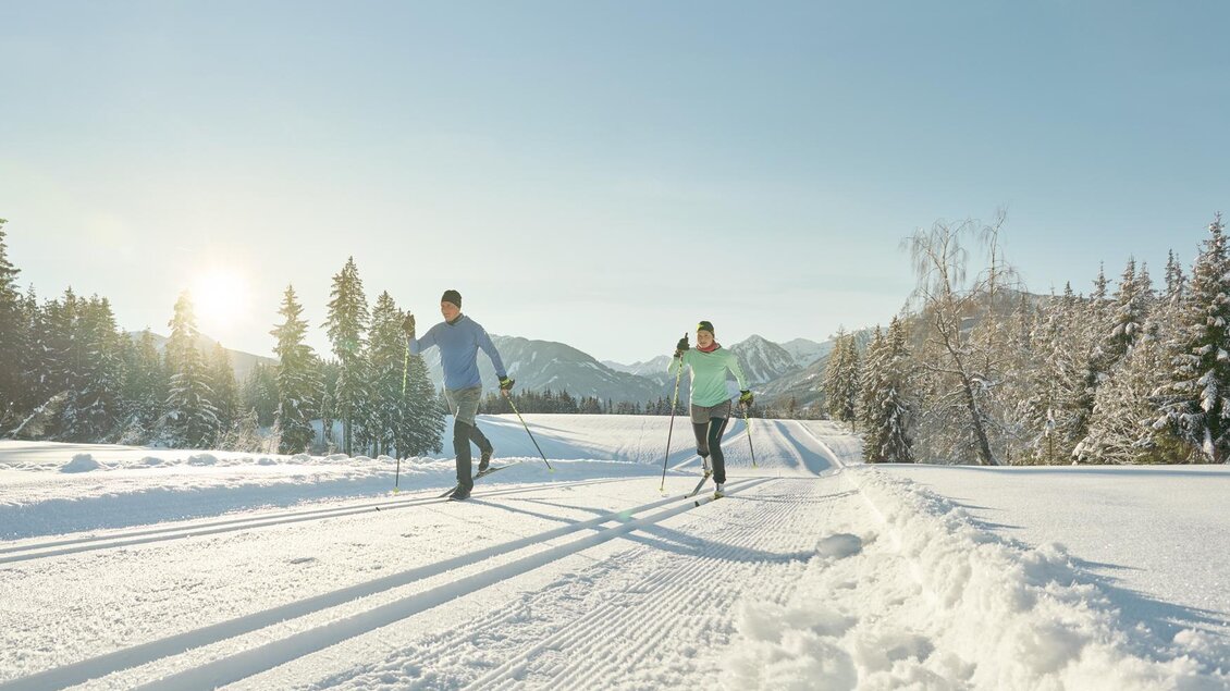Zwei Personen skaten im Schnee auf einer Langlaufloipe. Im Hintergrund sieht man verschneite Bäume und einen klaren Himmel.