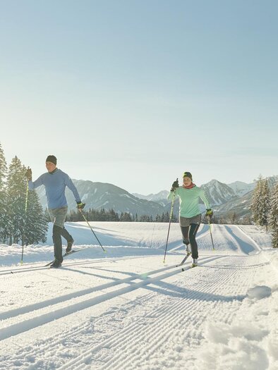 Zwei Personen skaten im Schnee auf einer Langlaufloipe. Im Hintergrund sieht man verschneite Bäume und einen klaren Himmel.