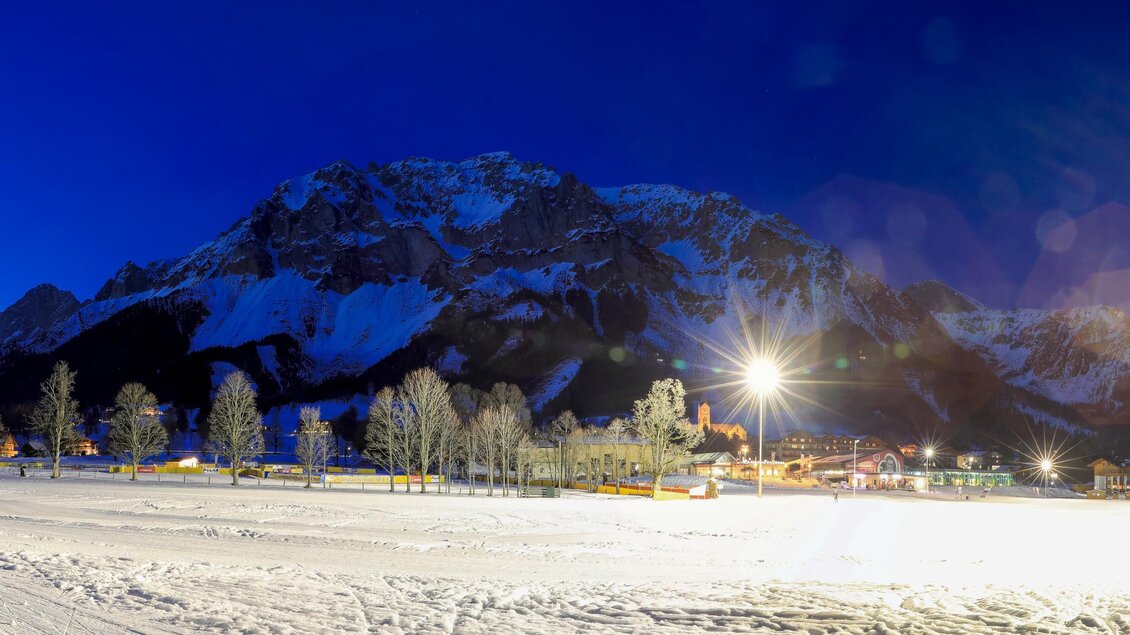 Eine verschneite Landschaft bei Nacht, umgeben von majestätischen Bergen. Strahlende Lichter erhellen die Umgebung und schaffen eine magische Atmosphäre.