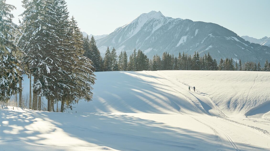 Eine winterliche Landschaft mit schneebedeckten Feldern und hohen Bergen im Hintergrund. Zwei Personen wandern durch die verschneite Umgebung. | © Peter Burgstaller