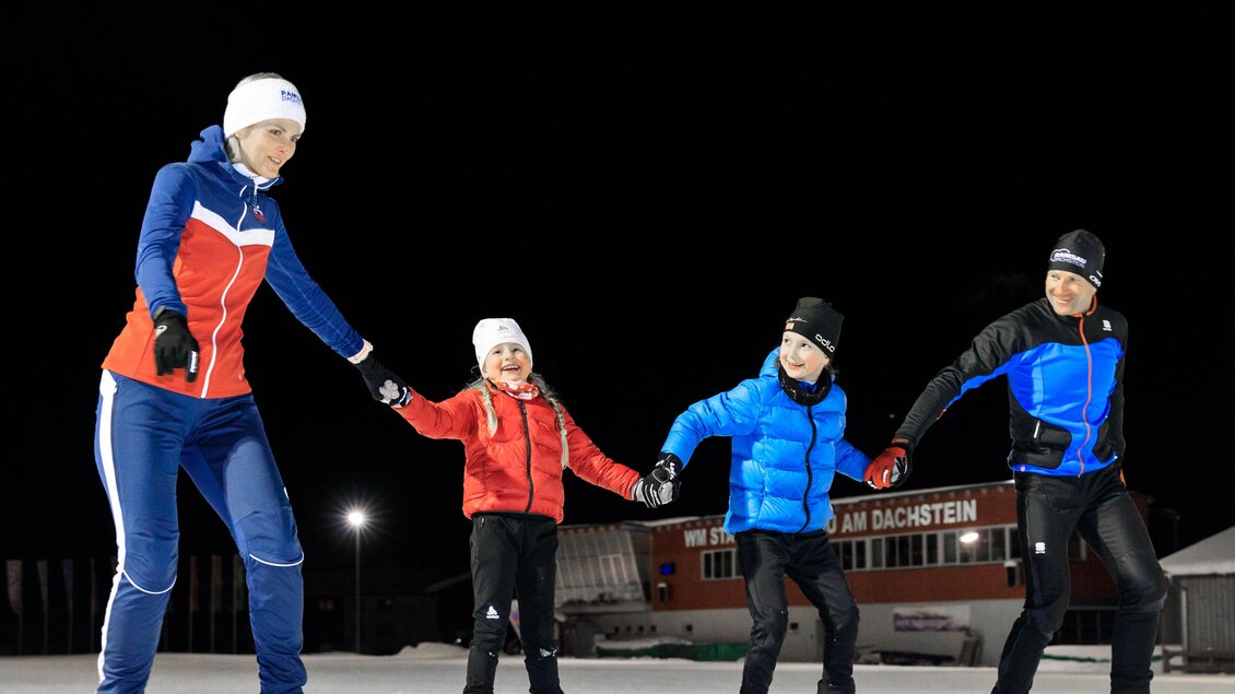 Eine Gruppe von vier Personen skiert abends auf einer beleuchteten Piste. Sie halten sich an den Händen und lachen zusammen.