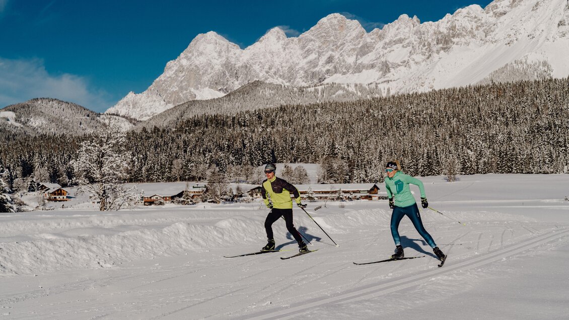 Zwei Skifahrer gleiten über eine schneebedeckte Landschaft, umgeben von majestätischen Bergen. Der klare Himmel und die dichten Wälder schaffen eine wunderschöne Winteratmosphäre. | © Christine Höflehner