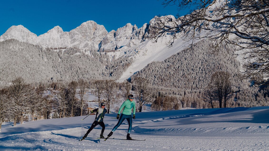 Zwei Skifahrer gleiten durch eine verschneite Landschaft. Im Hintergrund sind majestätische Berge und ein klarer blauer Himmel zu sehen. | © Christine Höflehner
