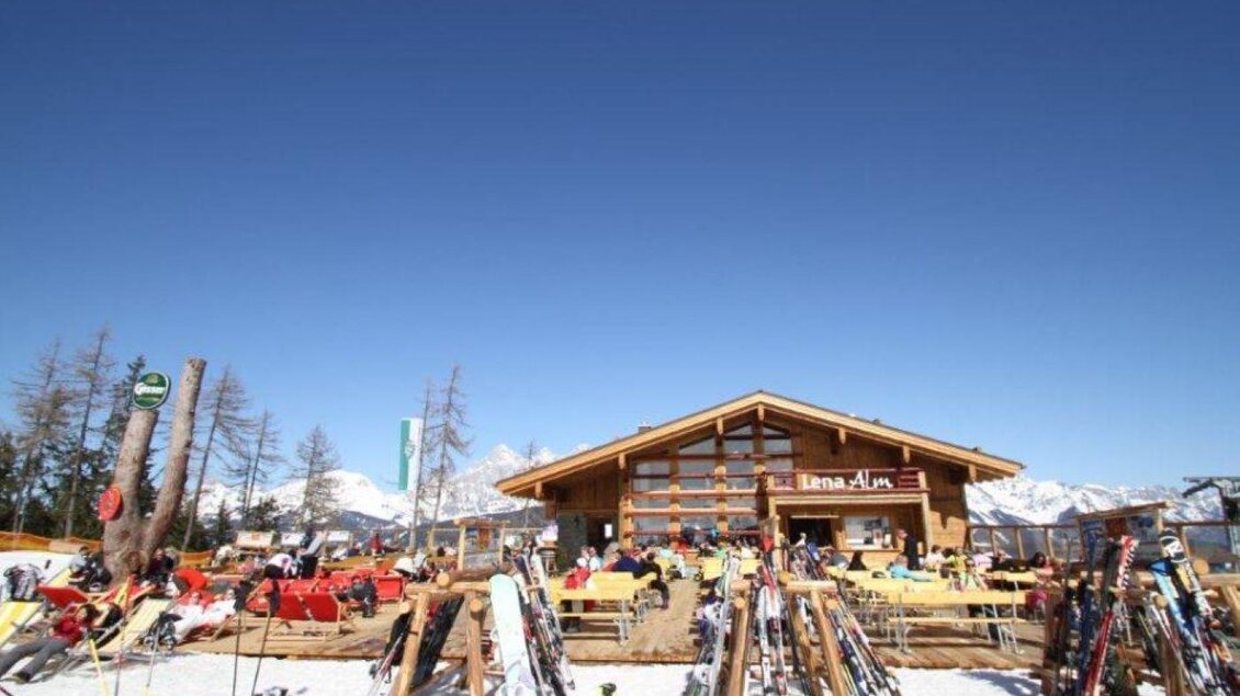 Ein gemütliches Skihütte in den Bergen mit einem klaren blauen Himmel. Vor der Hütte stehen zahlreiche Skier und gemütliche Sitzgelegenheiten. | © Lena-Alm