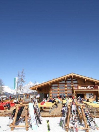 Ein gemütliches Skihütte in den Bergen mit einem klaren blauen Himmel. Vor der Hütte stehen zahlreiche Skier und gemütliche Sitzgelegenheiten. | © Lena-Alm