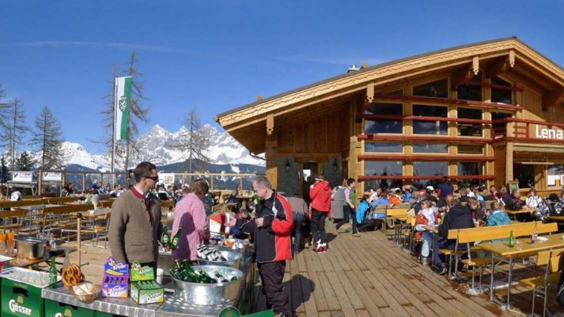 Eine alpine Berghütte mit großer Terrasse und vielen Gästen. Im Hintergrund sind schneebedeckte Berge und blauer Himmel zu sehen. | © Lena-Alm