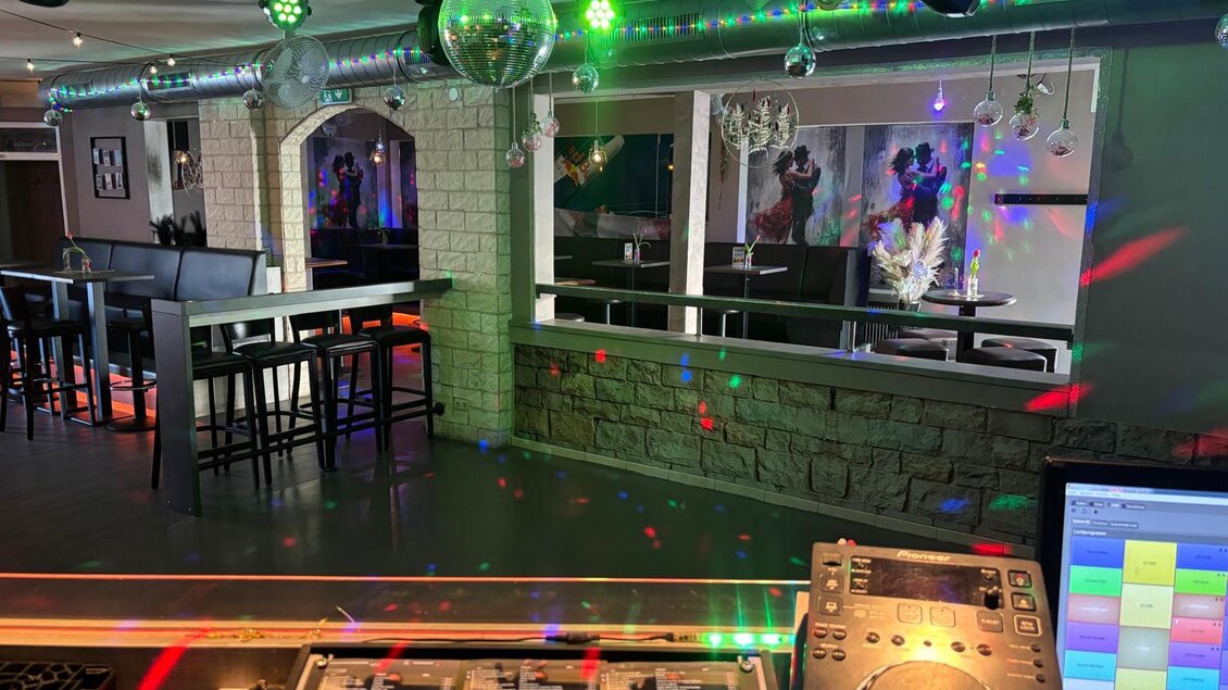 Eine moderne Bar mit bunten Lichtstrahlen und einer DJ-Konsole im Vordergrund. Im Hintergrund sind Tische und Stühle in einer stilvollen Einrichtung zu sehen. | © Let's Dance/Rainer Kohlhauser