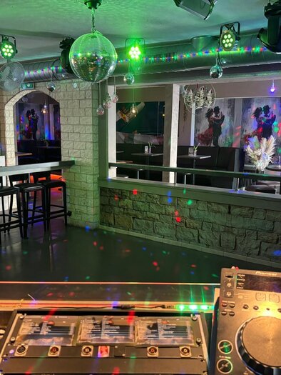 Eine moderne Bar mit bunten Lichtstrahlen und einer DJ-Konsole im Vordergrund. Im Hintergrund sind Tische und Stühle in einer stilvollen Einrichtung zu sehen. | © Let's Dance/Rainer Kohlhauser