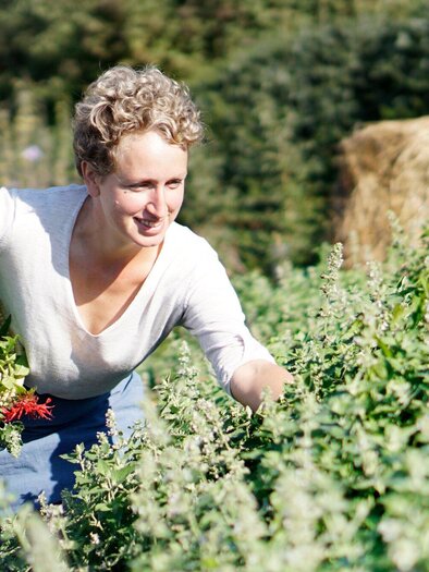 A woman is harvesting fresh herbs in a garden. In the background, hay bales and green plants can be seen. | © Sabine Lienhart