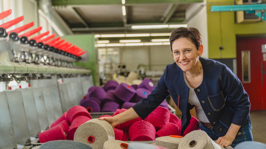 Eine Frau lächelt und steht in einer Fabrik neben großen Rollen von buntem Garn. Im Hintergrund sind Maschinen zu sehen, die zur Textilproduktion verwendet werden. | © Lodenmanufaktur Steiner1888 | Fotografin Christine Höflehner