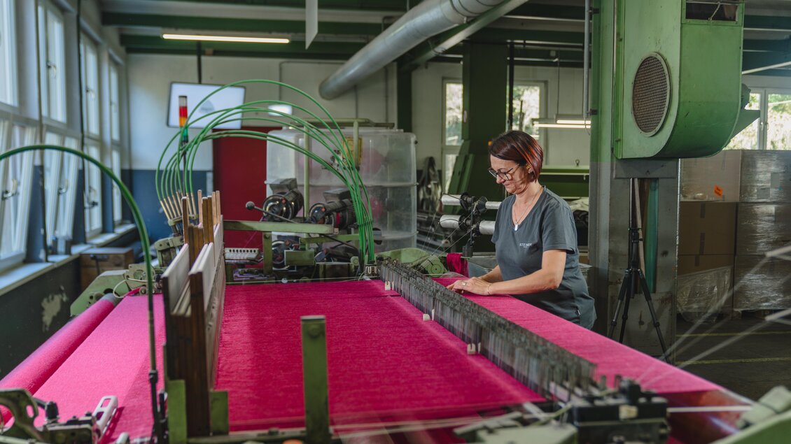 Eine Textilarbeiterin bedient eine Maschine in einer Fabrik. Der Fokus liegt auf einem leuchtend pinken Stoff, der hergestellt wird. | © Lodenmanufaktur Steiner1888 | Fotografin Christine Höflehner