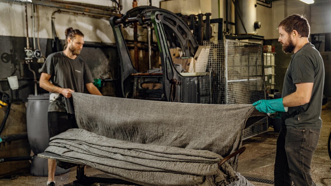 Zwei Männer heben eine große graue Decke in einer industriellen Umgebung. Sie tragen Arbeitshandschuhe und scheinen in einem Lager oder Werkstatt zu sein. | © Lodenmanufaktur Steiner1888 | Fotografin Christine Höflehner