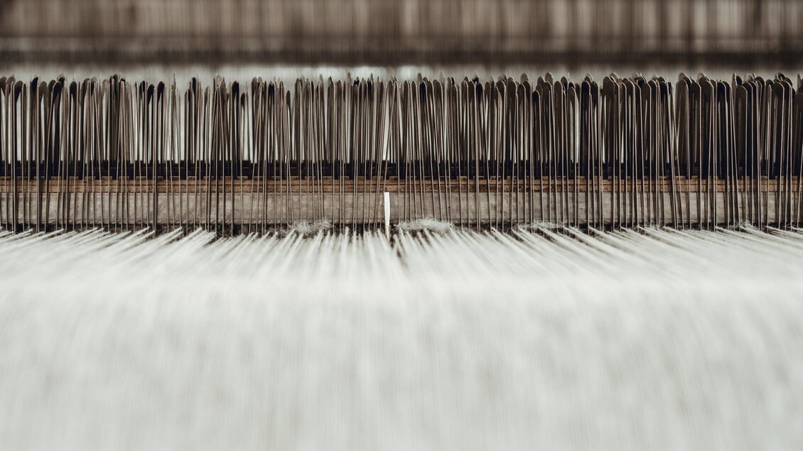 Eine Maschinenansicht mit vielen feinen Fäden, die in einem Textilproduktionsprozess angeordnet sind. Die Oberfläche erscheint glatt und strukturiert. | © © 2022 Alwin Strasser - strasserconcept