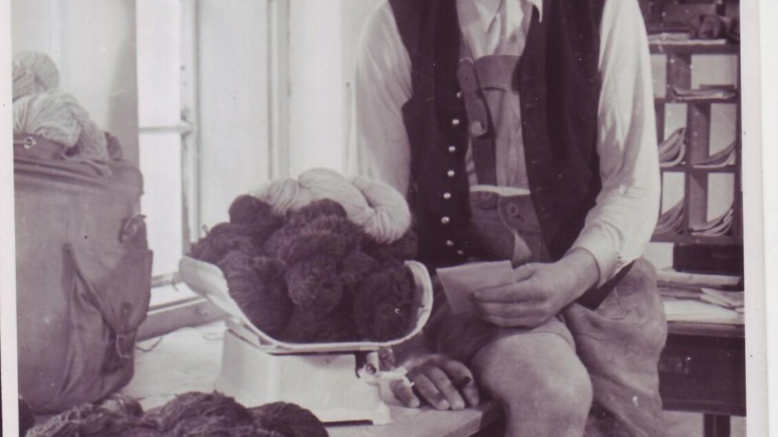Ein Mann in traditioneller Kleidung sitzt an einem Tisch, auf dem Wolle liegt. Er schaut in die Kamera und scheint nachdenklich zu sein. | © Lodenwalker Ramsau