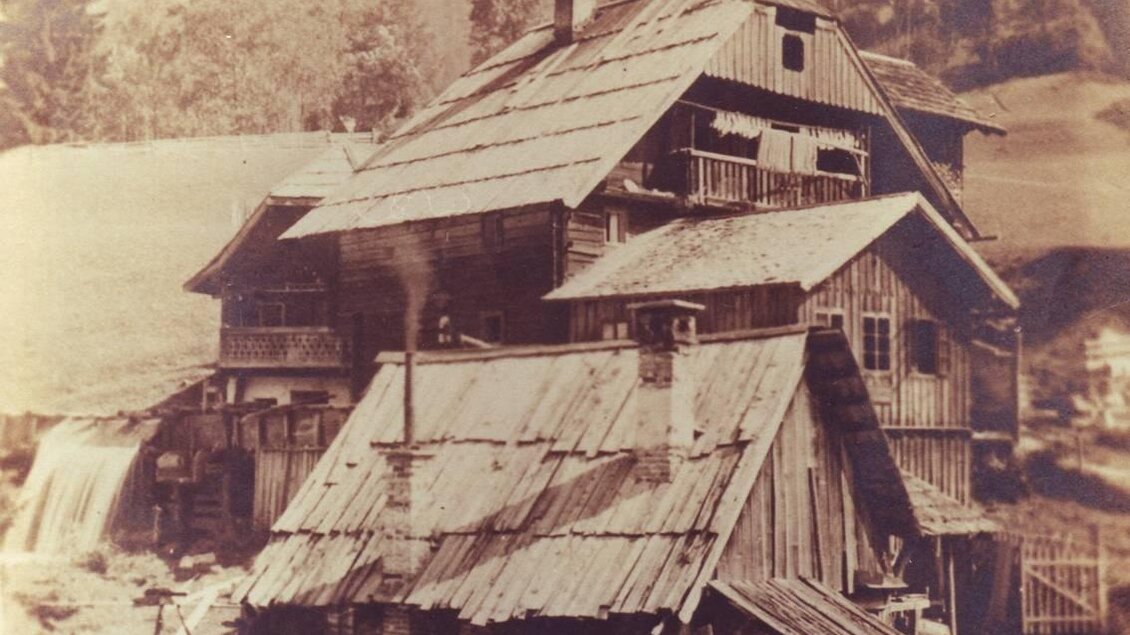 Ein historisches Gebäude mit einem rustikalen Dach und mehreren Stockwerken, umgeben von einer grünen Landschaft. Im Hintergrund sind Berge zu sehen. | © Lodenwalker Ramsau
