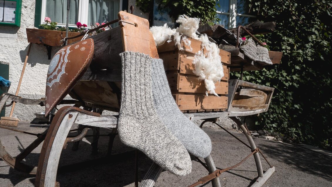 Ein alter Schlitten mit Holzkisten und Wollmaterialien steht vor einem Gebäude. Graue Socken hängen dekorativ am Schlitten. | © © 2021 Alwin Strasser - strasserconcept