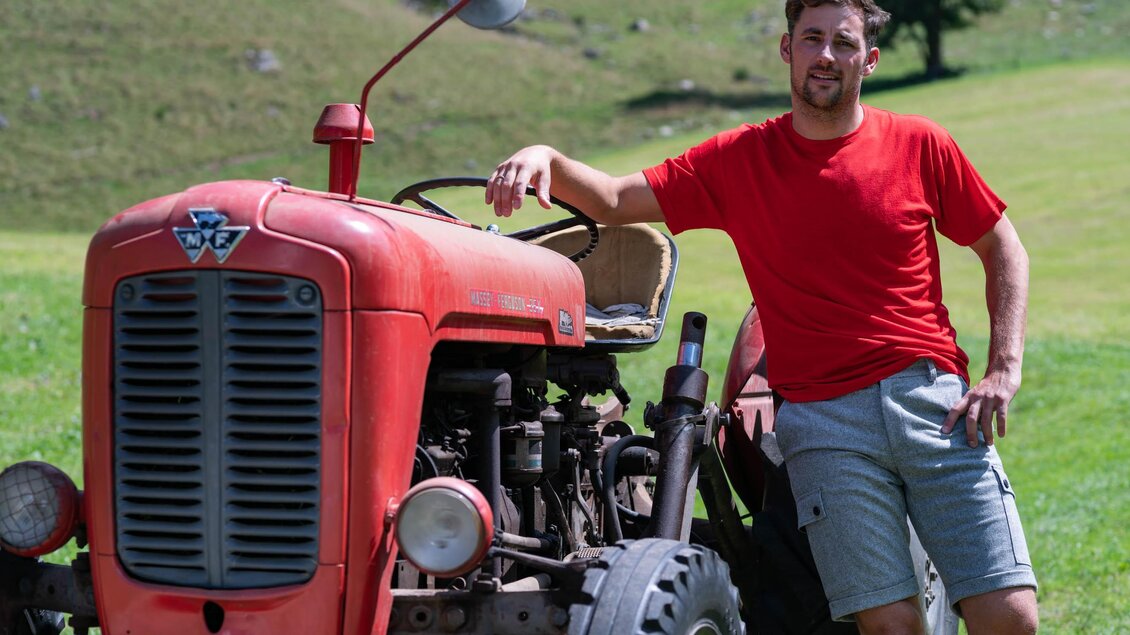 Ein Mann in einem roten T-Shirt steht neben einem alten roten Traktor auf einer grünen Wiese. Im Hintergrund sind sanfte Hügel zu sehen. | © © 2022 Andreas Pilz - KARAKORUM