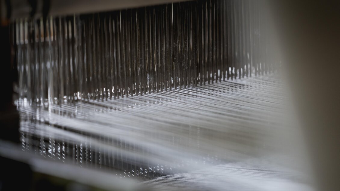 Ein detaillierter Blick auf eine Webmaschine mit vielen dünnen Fäden. Die reflektierende Oberfläche zeigt den industriellen Prozess der Textilherstellung. | © © 2021 Alwin Strasser - strasserconcept