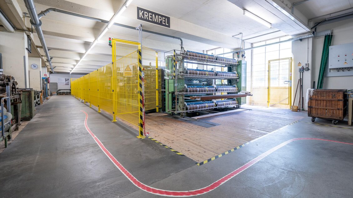 Eine helle Industriehalle mit Maschinen in einem Produktionsbereich. Gelbe Sicherheitsbarrieren umgeben die Arbeitsmaschinen.