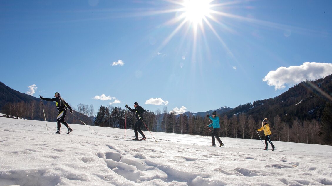 Eine Gruppe von vier Personen wandert über eine schneebedeckte Fläche im Winter. Die Sonne strahlt vom blauen Himmel und die Berge sind im Hintergrund sichtbar. | © Eva Rossböck