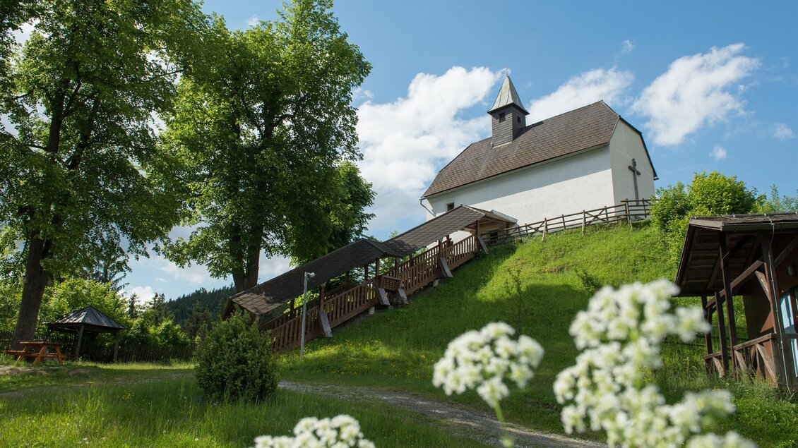 Eine kleine Kirche steht auf einem Hügel, umgeben von Bäumen und einer grünen Wiese. Der Himmel ist blau mit wenigen Wolken. | © Peter Wagner