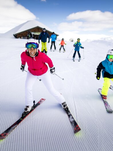 A group of skiers is skiing down a snow-covered slope. In the background, there is an Alpine scene with a ski hut visible. | © TVB Ausseerland Salzkammergut / Tom Lamm