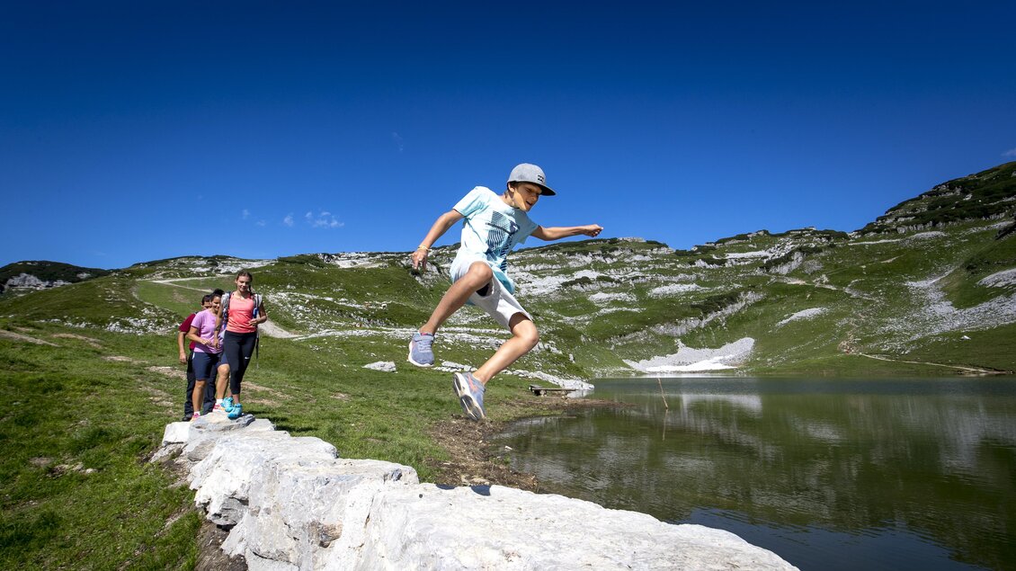 <p>Ein Junge springt über einen Felsenmauer am Rande des Augstsees. Im Hintergrund sind weitere Personen zu sehen, die dem Jungen nachgehen, umgeben von deb grünen Wiesen und Bergen am Loser.</p> | © TVB Ausseerland-Salzkammergut/Tom Lamm