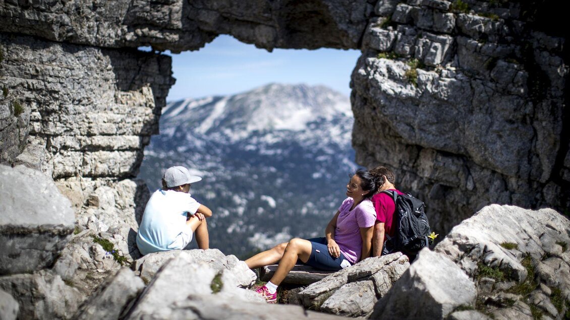<p>Über die Panoramastraße kann bis zum Parkplatz bei der Loseralm gefahren werden, von wo der Weg zum Loserfenster nicht mehr all zu weit ist. Drei Wanderer sitzen hier vor dem Loserfenster und genießen die Aussicht durch den natürlichen Felsbogen.</p> | © TVB Ausseerland - Salzkammergut-Tom Lamm