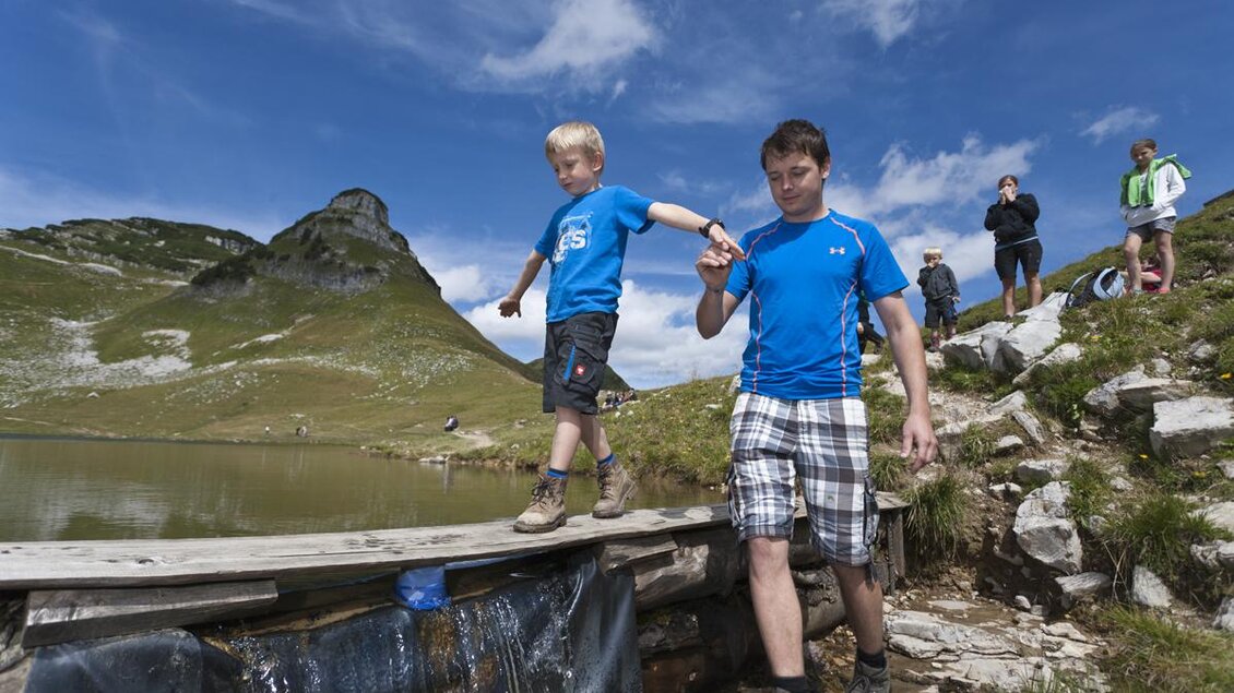 <p>Ein Junge balanciert über eine Holzbrücke und wird von einem Mann an der Hand gehalten. Unter ihm fließt das Wasser vom Augstsee. Im Hintergrund sind weitere Personen und auch der Atterkogel zu sehen.</p> | © Loser Bergbahnen Altaussee/Leo Himsl