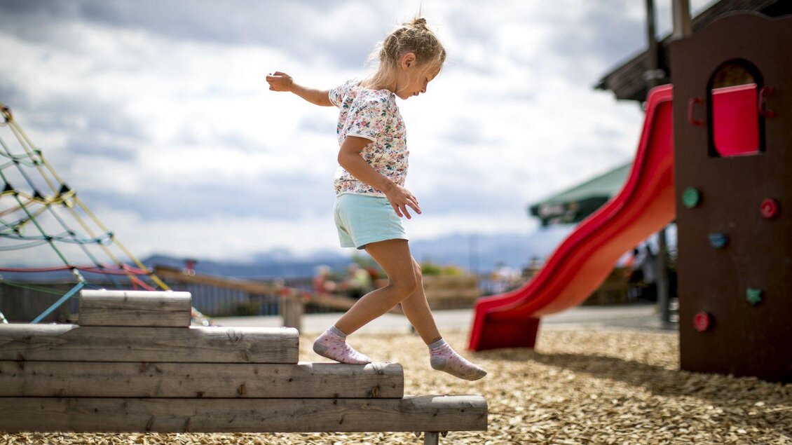 <p>Ein Mädchen balanciert spielerisch über einen Holzstamm auf dem Spielplatz neben der Loseralm. Im Hintergrund sind eine Rutsche und ein Klettergerüst sowie weitere Berge des Ausseerlandes zu sehen.</p> | © TVB Ausseerland - Salzkammergut-Tom Lamm