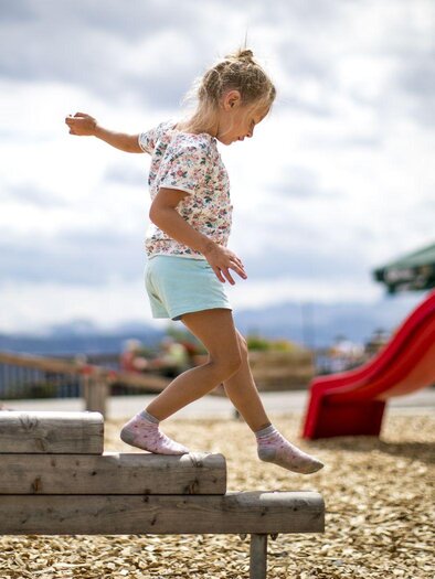 A girl is balancing on a wooden element in the playground. In the background, a slide and a climbing structure can be seen. | © TVB Ausseerland - Salzkammergut-Tom Lamm