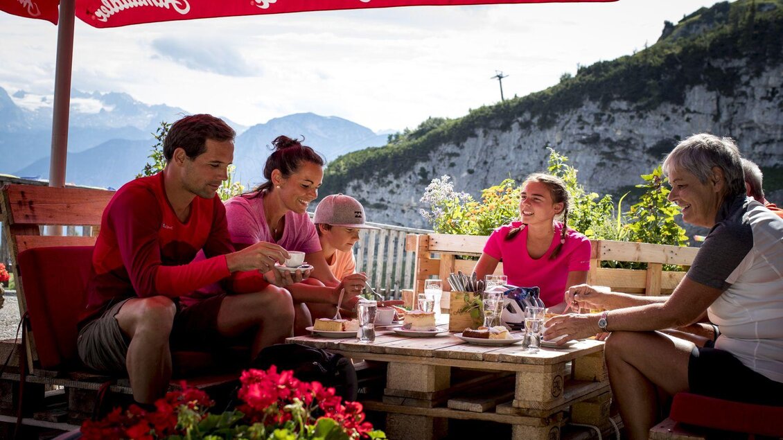 Eine fröhliche Gruppe von fünf Personen sitzt an einem Tisch im Freien und genießt eine Mahlzeit. Im Hintergrund sind Berge und grüne Landschaften zu sehen. | © TVB Ausseerland - Salzkammergut/Tom Lamm