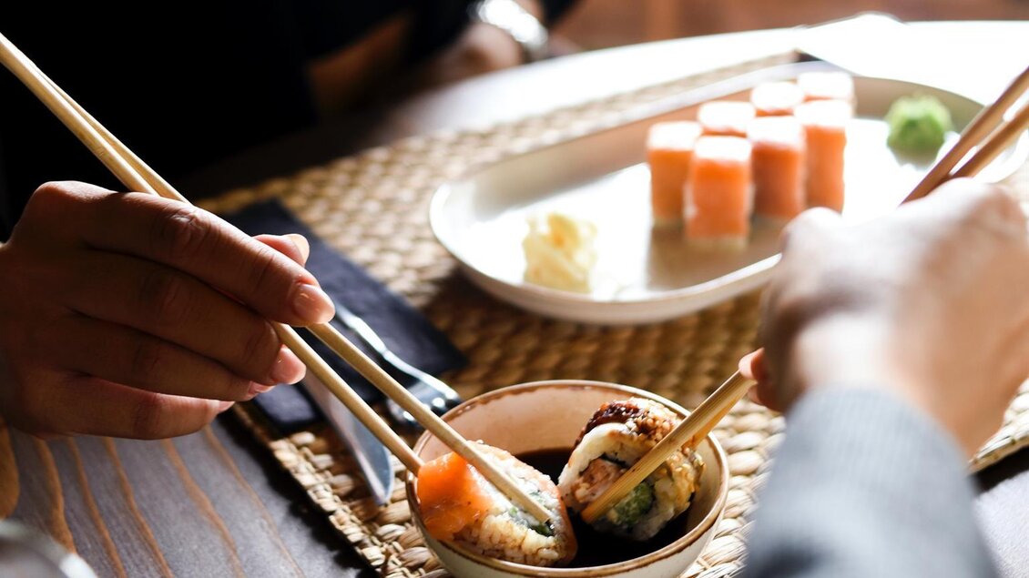 Zwei Hände, die Sushi mit Essstäbchen essen. Im Hintergrund sind verschiedene Sushi-Gerichte und eine kleine Schale mit Sojasauce zu sehen. | © Elif Tekkaya
