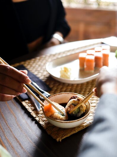 Zwei Hände, die Sushi mit Essstäbchen essen. Im Hintergrund sind verschiedene Sushi-Gerichte und eine kleine Schale mit Sojasauce zu sehen. | © Elif Tekkaya