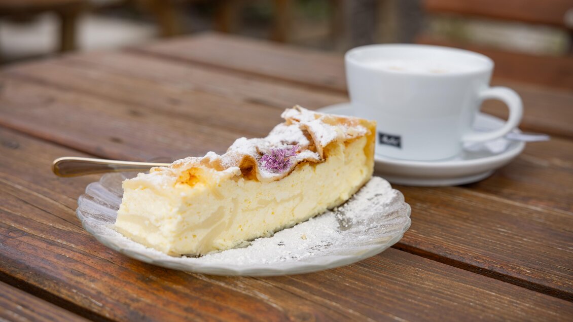 Ein Stück Käsekuchen auf einem Teller mit Puderzucker und eine Tasse Kaffee auf einem Holztisch. Die entspannte Atmosphäre lädt zum Genießen ein. | © 5komma5sinne