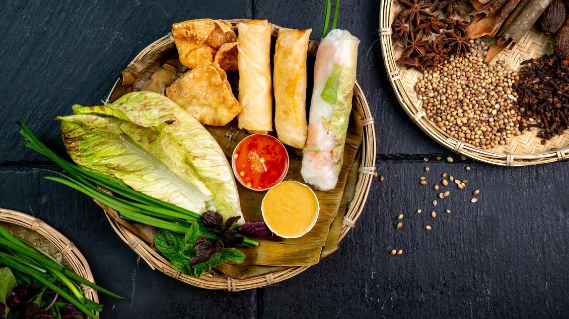 Frühlingsrollen, Wan-Tans, Reispapierrolle, Blattsalat, Kräuter und Dips auf Bambusteller angerichtet. | © Má Kitchen