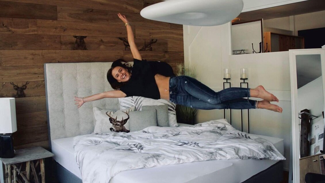<p>Eine Frau springt fröhlich auf ein Bett in einem modern eingerichteten Schlafzimmer von Möbel Peer in Bad Aussee. Die Wände sind aus Holz und die Dekoration ist schlicht und elegant.</p> | © Möbel Peer_Sabine Stöckl