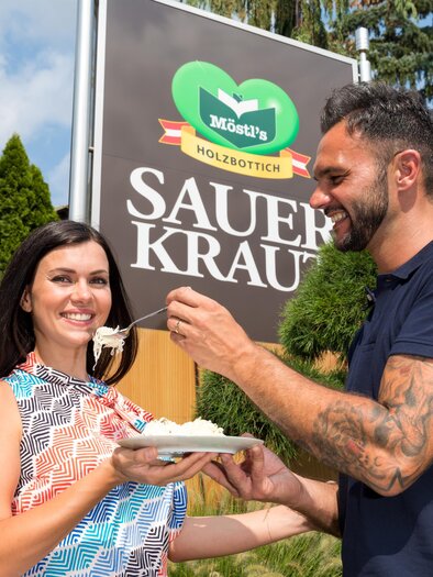 A man is feeding a woman with a spoonful of sauerkraut in front of a sign. In the background, green trees can be seen. | © Region Graz - Harry Schiffer
