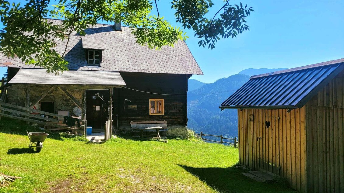 Ein traditionelles Holzhaus inmitten einer grünen Wiese. Im Hintergrund sind Berge und ein blauer Himmel zu sehen. | © Müller